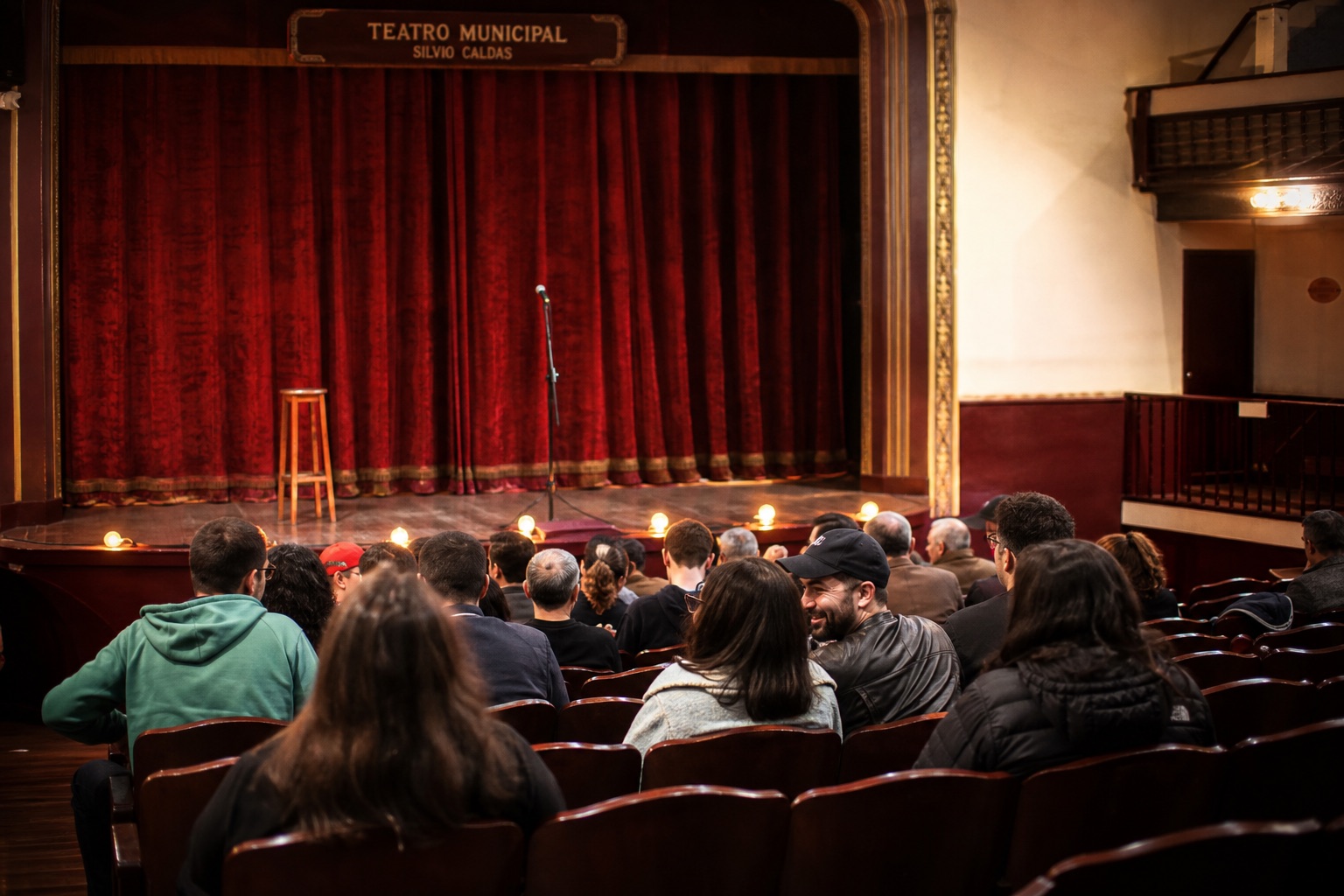 Interior de un teatro histórico antes de la función: telón de terciopelo rojo, luces de candilejas y un taburete con micrófono en el escenario